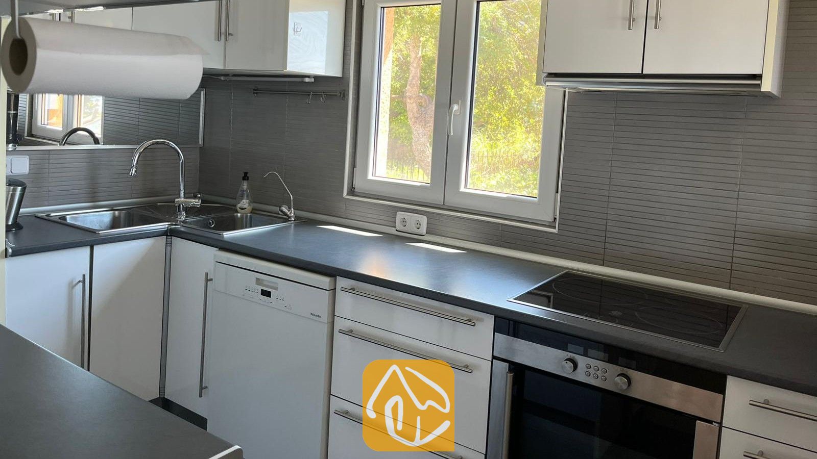 Vakantievilla's Costa Brava Spanje - Villa VistaMar - Moderne keuken met witte kasten, donkere aanrechtbladen, een ingebouwde oven en een dubbele spoelbak onder een raam.