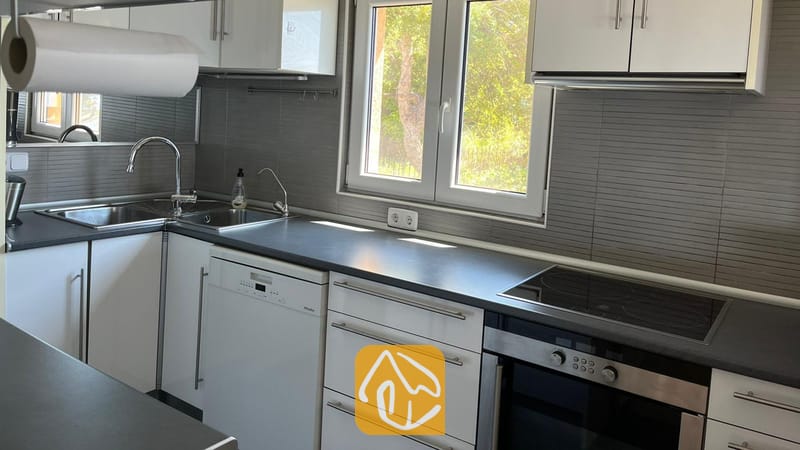 Vakantievilla's Costa Brava Spanje - Villa VistaMar - Moderne keuken met witte kasten, donkere aanrechtbladen, een ingebouwde oven en een dubbele spoelbak onder een raam.