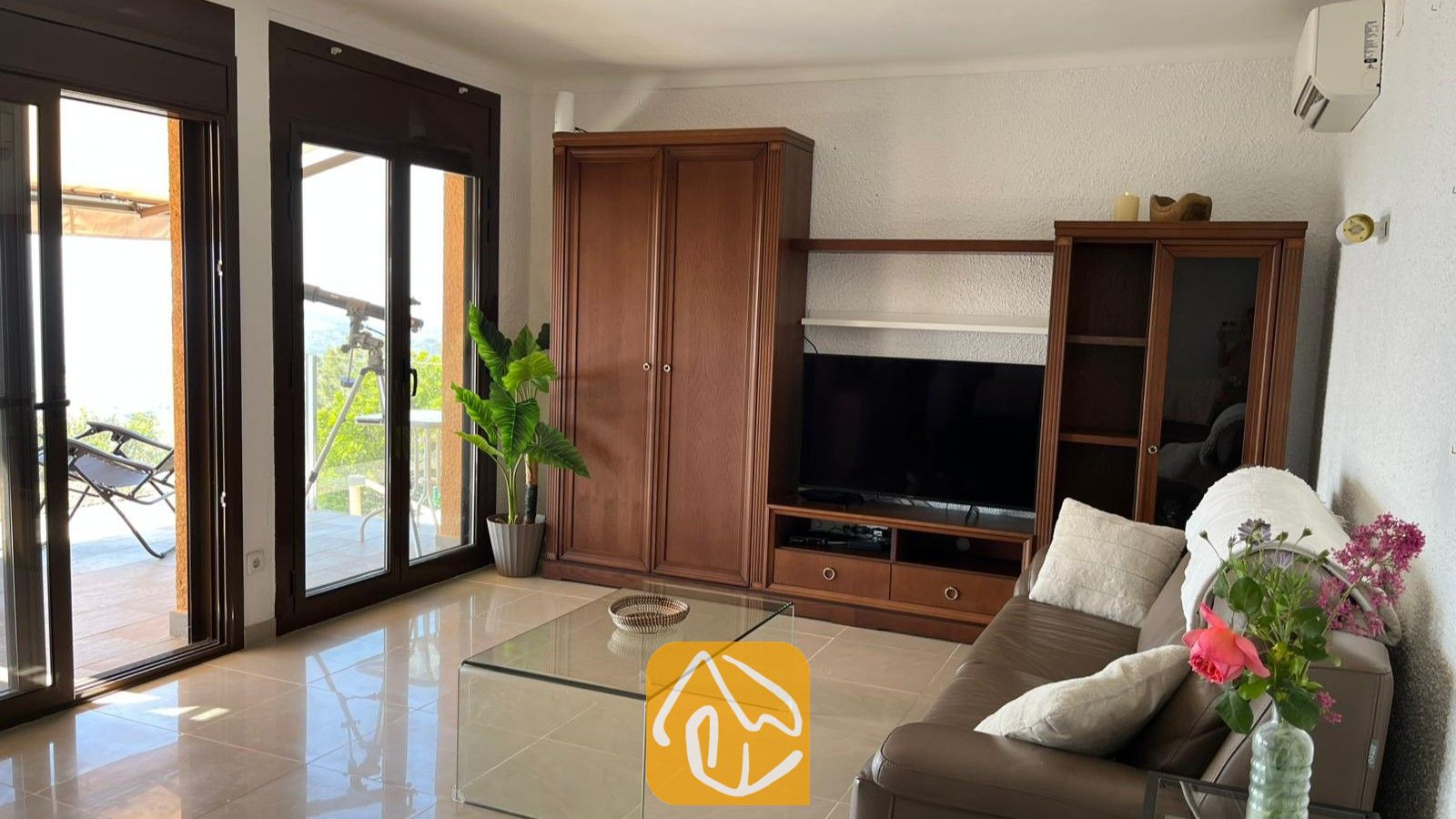Vakantievilla's Costa Brava Spanje - Villa VistaMar - Woonkamer voorzien van een donkerhouten meubel voor apparatuur, een glazen salontafel en glazen schuifdeuren die uitkomen op een zonnig terras bij Villa VistaMar.