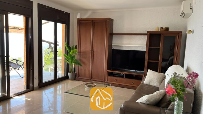 Vakantievilla's Costa Brava Spanje - Villa VistaMar - Woonkamer voorzien van een donkerhouten meubel voor apparatuur, een glazen salontafel en glazen schuifdeuren die uitkomen op een zonnig terras bij Villa VistaMar.