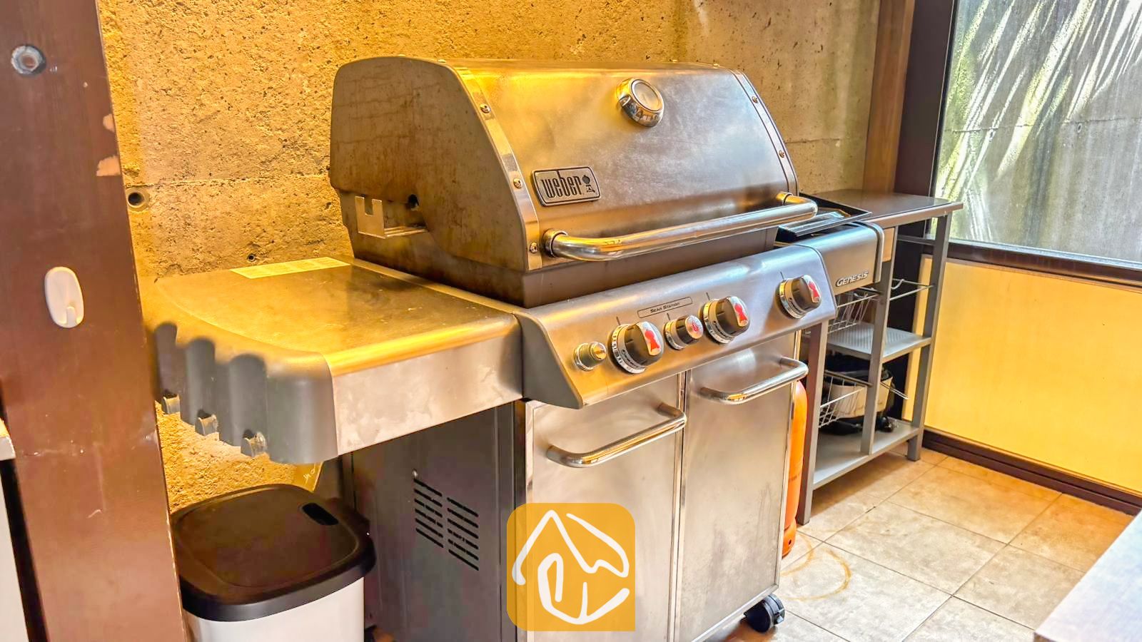 Vakantievilla's Costa Brava Spanje - Villa VistaMar - Een roestvrijstalen Weber gasbarbecue op een buitenterras met betegelde vloer en een wand van structuurbeton.