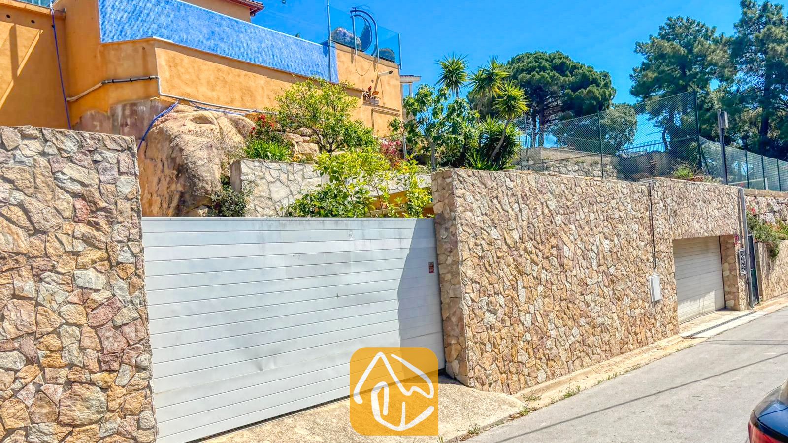 Vakantievilla's Costa Brava Spanje - Villa VistaMar - Buitenaanzicht van Villa VistaMar met een stenen erfbak, een witte schuifpoort van de garage en de geel-blauwe gevel van het huis tegen een heldere hemel.