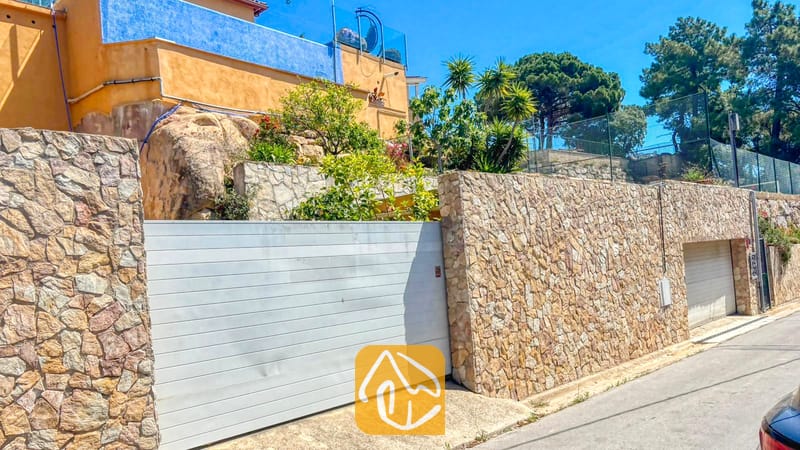 Villas de vacances Costa Brava Espagne - Villa VistaMar - Vue extérieure de la Villa VistaMar montrant un mur d'enceinte en pierre, un portail de garage coulissant blanc et la façade jaune et bleue de la maison sur fond de ciel dégagé.