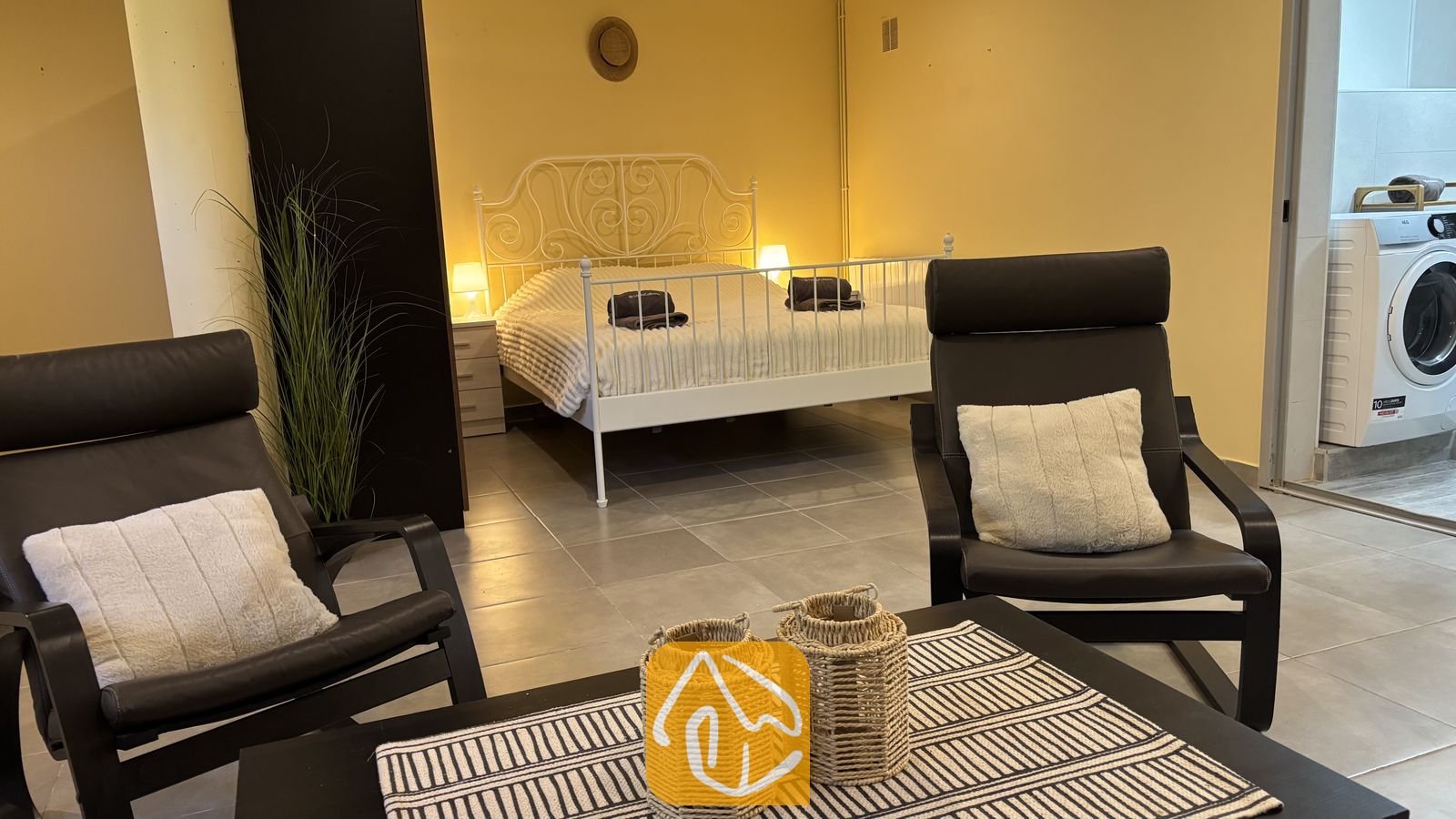 Vakantievilla's Costa Brava Spanje - Villa VistaMar - Slaapkamer met een tweepersoonsbed met een wit metalen frame, gele muren en op de voorgrond twee donkere fauteuils met witte kussens.