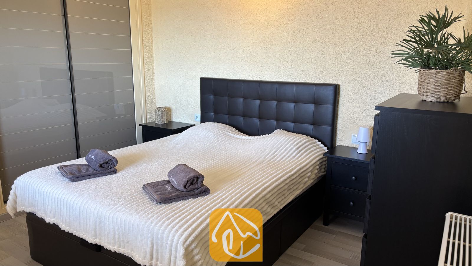Vakantievilla's Costa Brava Spanje - Villa VistaMar - Slaapkamer met een tweepersoonsbed voorzien van een donker gewatteerd hoofdbord, een witte textuursprei en opgevouwen grijze handdoeken bovenop, geflankeerd door twee kleine zwarte nachtkastjes.