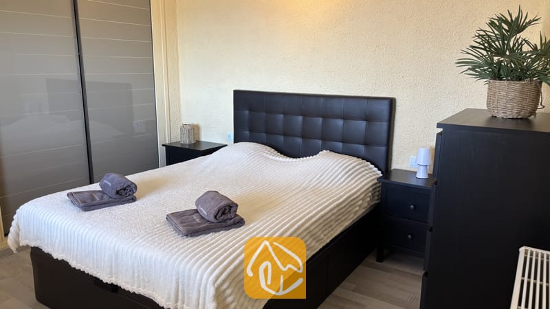 Villas de vacances Costa Brava Espagne - Villa VistaMar - Chambre avec un lit double doté d'une tête de lit matelassée foncée, d'une couverture blanche texturée et de serviettes grises pliées sur le dessus, encadrée par deux petites tables de chevet noires.