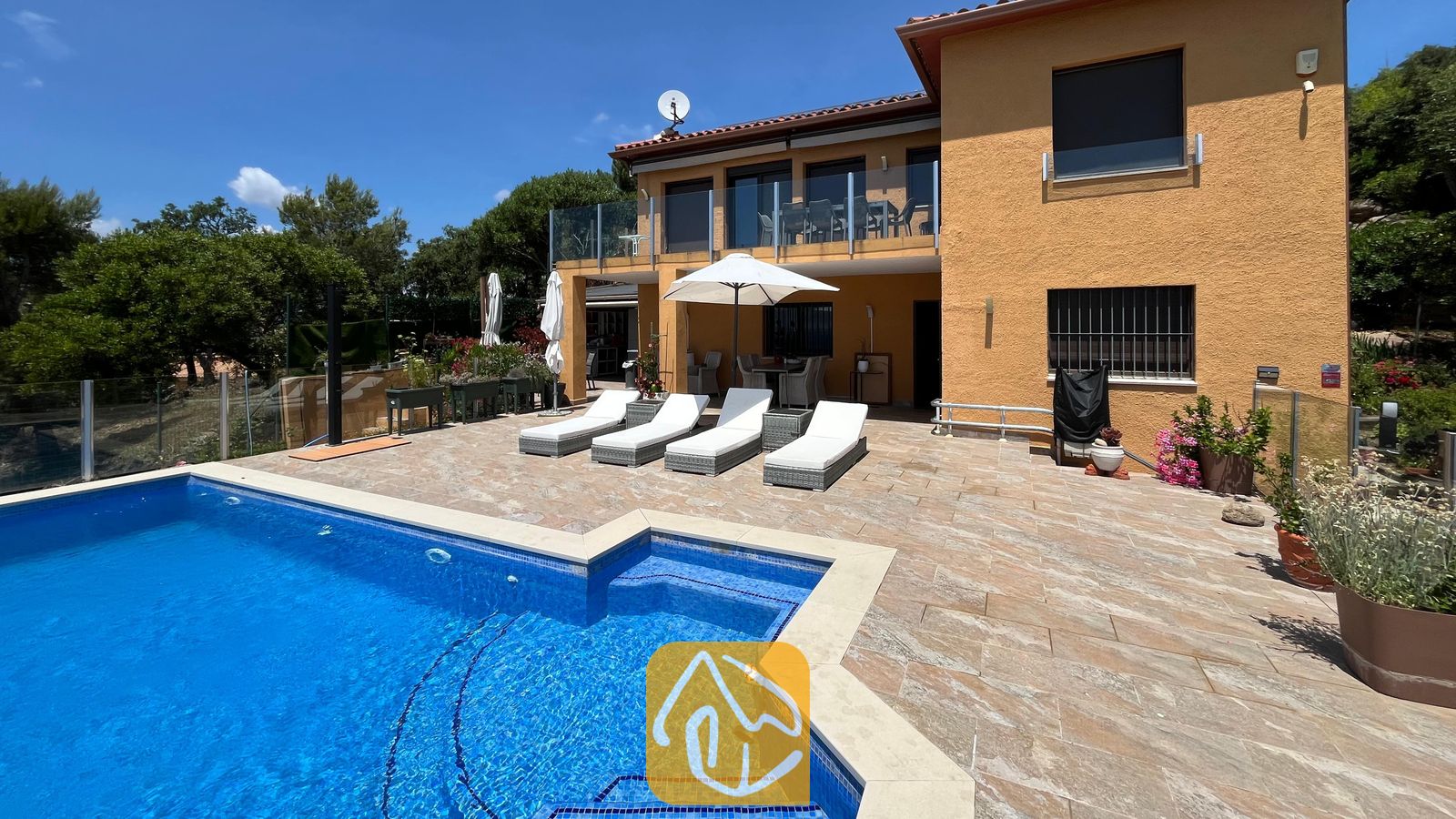 Vakantievilla's Costa Brava Spanje - Villa VistaMar - Buitenaanzicht van Villa VistaMar met een betegeld terras voorzien van vier witte ligstoelen, een parasol en een blauw betegeld zwembad tegen een gele stucwerk gevel.