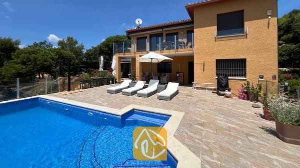 Vakantievilla's Costa Brava Spanje - Villa VistaMar - Buitenaanzicht van Villa VistaMar met een betegeld terras voorzien van vier witte ligstoelen, een parasol en een blauw betegeld zwembad tegen een gele stucwerk gevel.