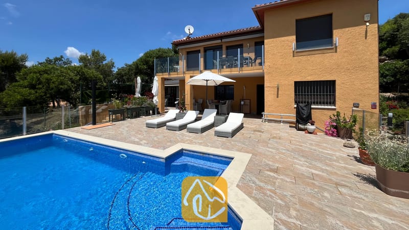 Vakantievilla's Costa Brava Spanje - Villa VistaMar - Buitenaanzicht van Villa VistaMar met een betegeld terras voorzien van vier witte ligstoelen, een parasol en een blauw betegeld zwembad tegen een gele stucwerk gevel.