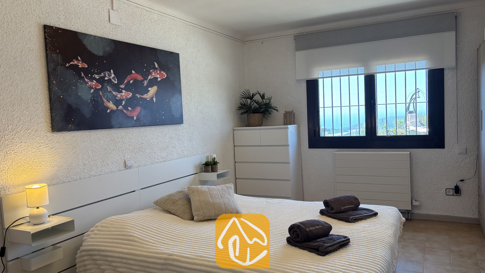 Vakantievilla's Costa Brava Spanje - Villa VistaMar - Slaapkamer voorzien van een tweepersoonsbed met opgevouwen donkere handdoeken, wit getextureerde muren en een groot schilderij van een koikarper boven het hoofdeinde bij Villa VistaMar.