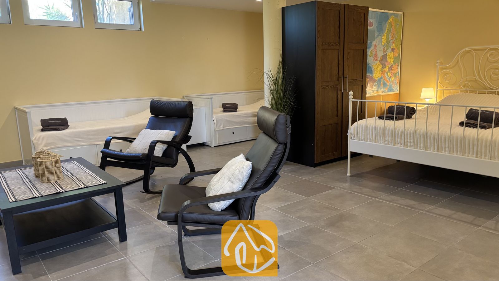 Vakantievilla's Costa Brava Spanje - Villa VistaMar - Ruim slaapgedeelte in Villa VistaMar, voorzien van een wit metalen framebed, donkere leren fauteuils en twee enkele daybeds tegen de gele wand.