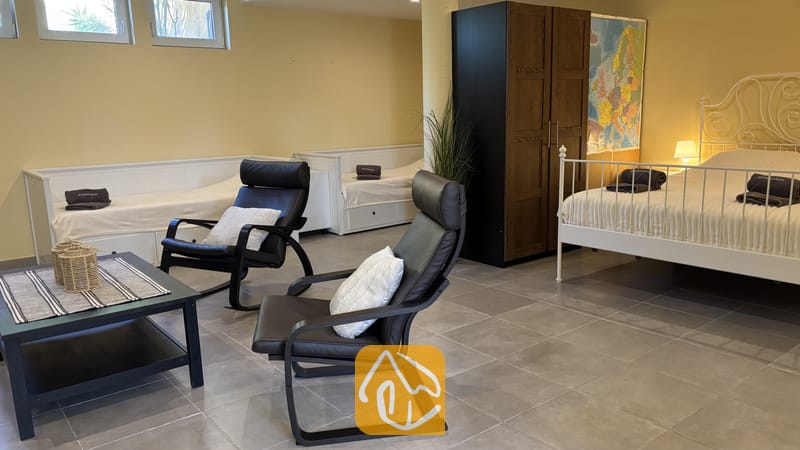 Villas de vacances Costa Brava Espagne - Villa VistaMar - Spacieuse chambre de la Villa VistaMar dotée d'un lit avec structure en métal blanc, de fauteuils en cuir foncé et de deux lits de repos individuels adossés au mur jaune.
