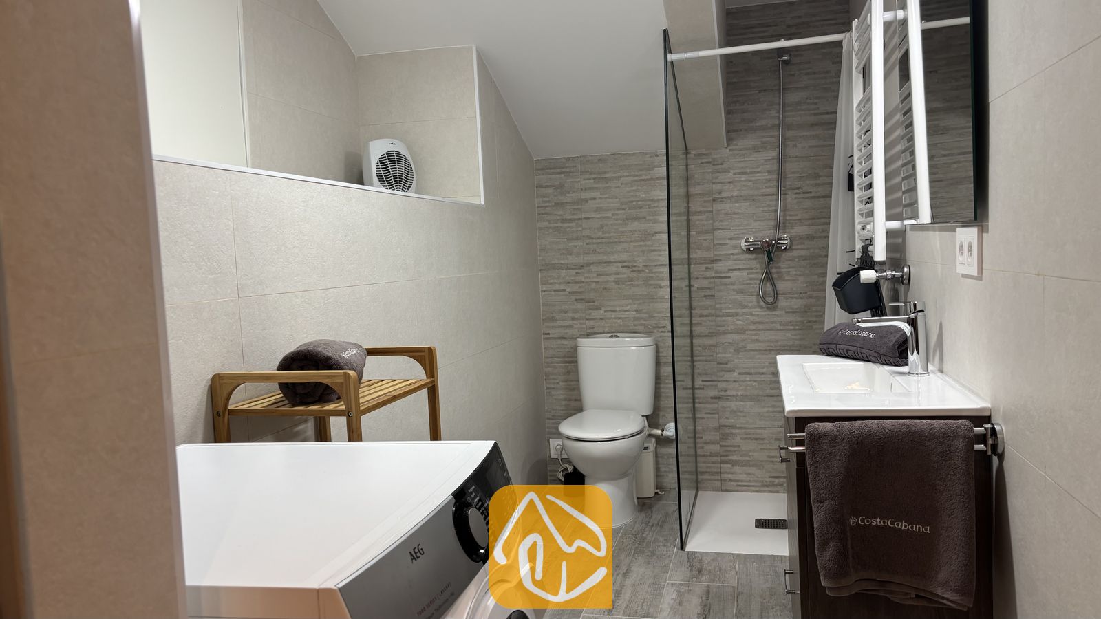 Vakantievilla's Costa Brava Spanje - Villa VistaMar - Moderne, betegelde badkamer met een witte wasmachine, een inloopdouche met glazen wand, toilet en houten planken met opgerolde grijze handdoeken.