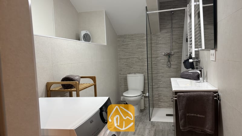 Villas de vacances Costa Brava Espagne - Villa VistaMar - Salle de bains moderne carrelée comprenant un lave-linge blanc, une douche à l'italienne avec paroi en verre, des toilettes et des étagères en bois accueillant des serviettes grises roulées.