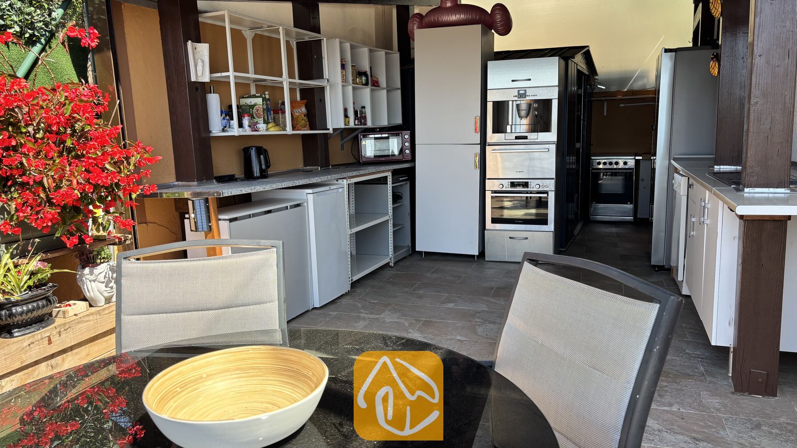 Vakantievilla's Costa Brava Spanje - Villa VistaMar - Buitenkeuken met witte kasten, ingebouwde roestvrijstalen apparatuur en op de voorgrond een eettafel met grijs mesh stoelen.