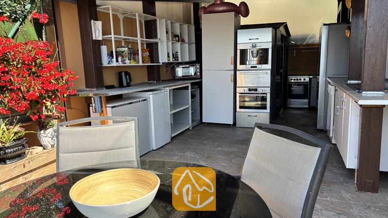 Villas de vacances Costa Brava Espagne - Villa VistaMar - Coin cuisine extérieur comprenant des placards blancs, des appareils encastrés en acier inoxydable et une table à manger avec des chaises en résine tressée grise au premier plan.