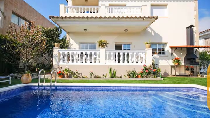 Vakantievilla's Costa Brava Spanje - Villa Mercedes Fenals - Privézwembad met helderblauw water voor de buitenkant van Villa Mercedes Fenals, voorzien van balkons met witte balustrades, een gazon en een stenen barbecue in de buitenlucht.