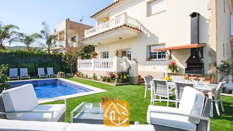 Vakantievilla's Costa Brava Spanje - Villa Mercedes Fenals - Buitenaanzicht van Villa Mercedes Fenals, met een zwembad, gazon, witte tuinstoelen en een ingebouwde buitenbarbecue tegen de crèmekleurige gevel.