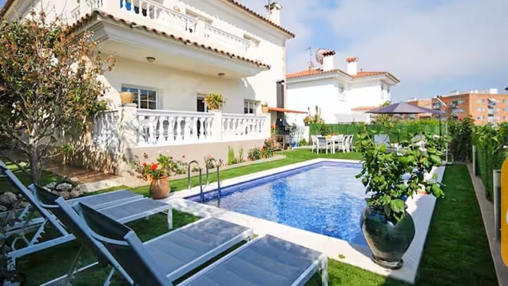 Vakantievilla's Costa Brava Spanje - Villa Mercedes Fenals - Buitenaanzicht van Villa Mercedes Fenals met een rechthoekig zwembad omringd door grijs ligbedden, een gazon, potplanten en de witte gevel van de villa met meerdere verdiepingen op de achtergrond.