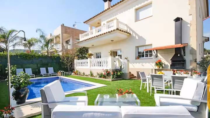 Vakantievilla's Costa Brava Spanje - Villa Mercedes Fenals - Buitenaanzicht van Villa Mercedes Fenals met een eigen zwembad, een groen gazon, witte 
tuinmeubelen en een aangebouwde barbecuehoek.