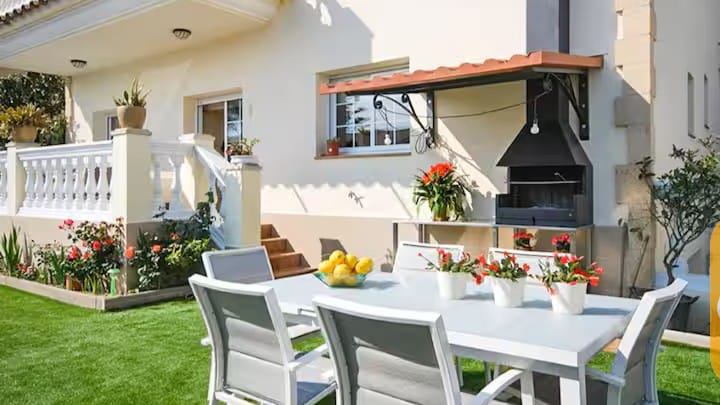 Vakantievilla's Costa Brava Spanje - Villa Mercedes Fenals - Buiteneteenhoek bij Villa Mercedes Fenals met een witte metalen tafel, bijpassende stoelen, rode potplanten en een ingebouwde barbecue tegen de buitenmuur.
