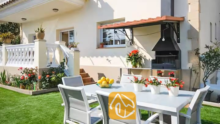 Vakantievilla's Costa Brava Spanje - Villa Mercedes Fenals - Buiteneteenhoek bij Villa Mercedes Fenals met een witte metalen tafel, bijpassende stoelen, rode potplanten en een ingebouwde barbecue tegen de buitenmuur.