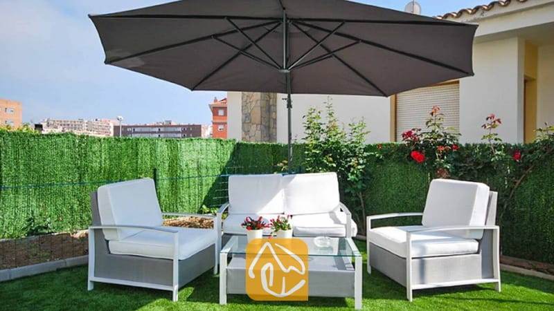 Vakantievilla's Costa Brava Spanje - Villa Mercedes Fenals - Loungehoek op het buitenterras van Villa Mercedes Fenals, voorzien van een grijze parasol, wit gekussende fauteuils, een bijpassende bank en een salontafel met glazen blad op kunstgras.