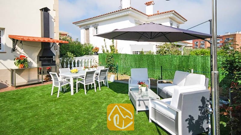 Vakantievilla's Costa Brava Spanje - Villa Mercedes Fenals - Eetgedeelte buiten en loungesets op het kunstgrasveld bij Villa Mercedes Fenals, voorzien van een zwarte stenen barbecue en een grote grijze parasol.