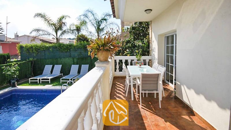 Vakantievilla's Costa Brava Spanje - Villa Mercedes Fenals - Balkon van Villa Mercedes Fenals met een eettafel en stoelen buiten, uitkijkend over het zwembad en de palmbomen beneden.