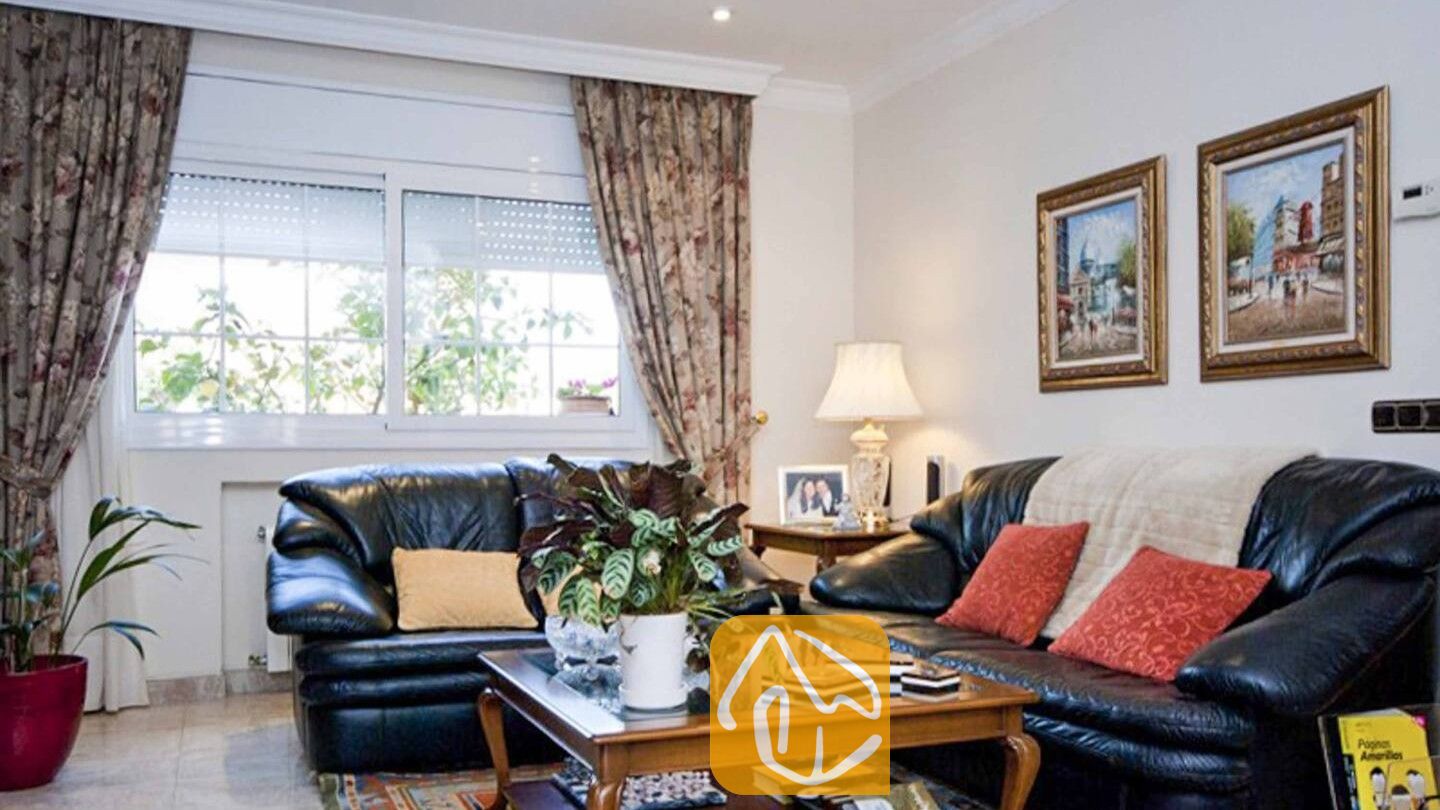 Vakantievilla's Costa Brava Spanje - Villa Mercedes Fenals - Woonkamer met twee zwarte leren banken voorzien van rode en gele sierkussens, bloemengordijnen, een houten salontafel en ingelijste schilderijen aan de muur.