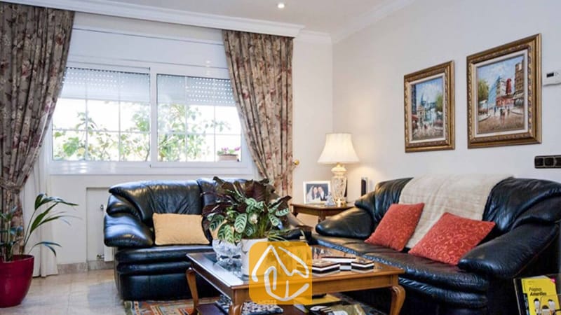 Ferienhäuser Costa Brava Spanien - Villa Mercedes Fenals - Wohnbereich mit zwei schwarzen Ledersofas mit roten und gelben Zierkissen, floralen Vorhängen, einem Couchtisch aus Holz und gerahmten Gemälden an der Wand.