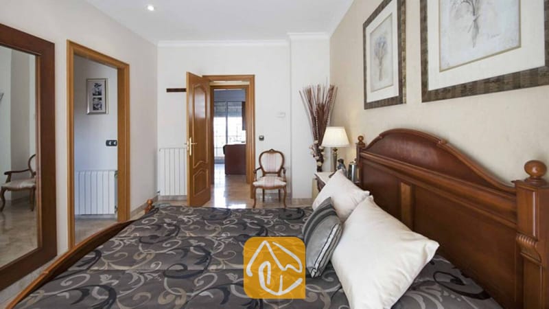 Ferienhäuser Costa Brava Spanien - Villa Mercedes Fenals - Schlafzimmer mit einem dunklen Holzkopfteil, gemusterter Bettwäsche, weißen Kissen und gerahmten Kunstwerken an beigefarbenen Wänden.