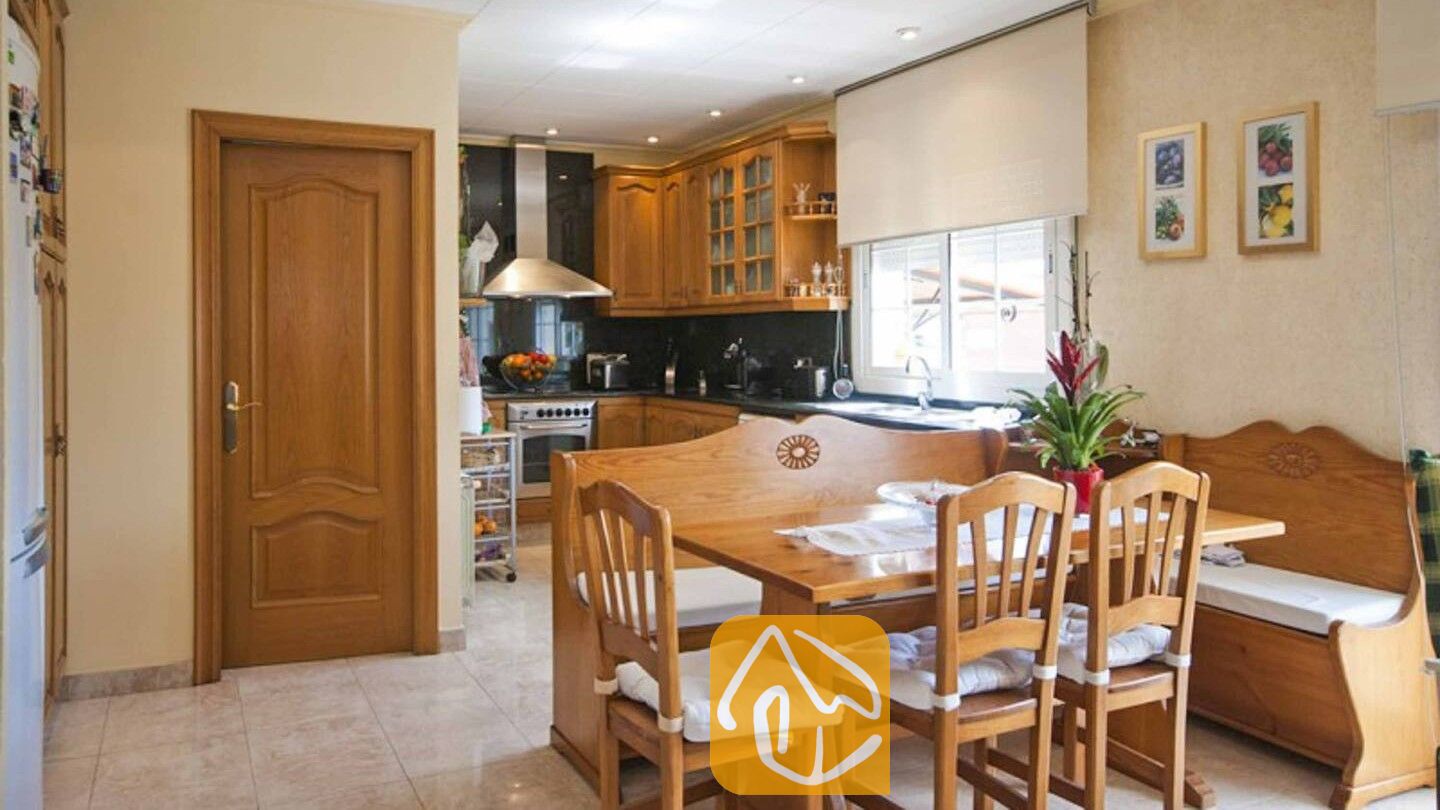 Vakantievilla's Costa Brava Spanje - Villa Mercedes Fenals - Houten eettafel met stoelen en een bankje in Villa Mercedes Fenals, met op de achtergrond een keuken met houten kasten, tegelvloeren en ingelijste muurkunst.