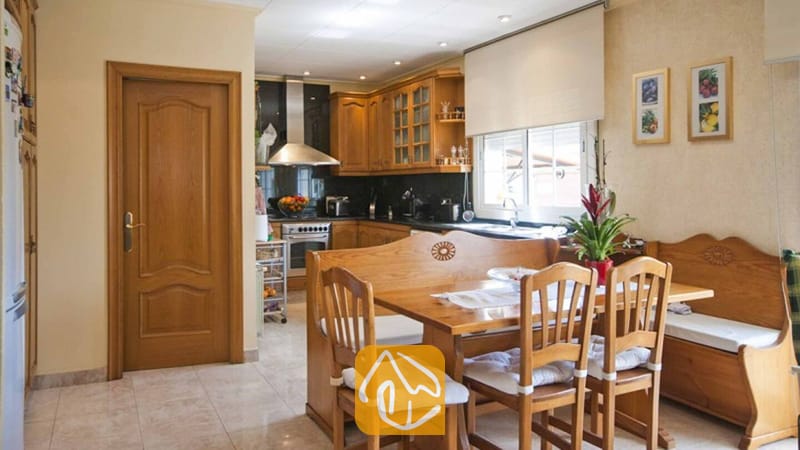 Ferienhäuser Costa Brava Spanien - Villa Mercedes Fenals - Massivholzesstisch mit Stühlen und Sitzbank im Inneren der Villa Mercedes Fenals, vor der Kulisse einer Küche mit Holzschränken, Fliesenböden und gerahmter Wandkunst.