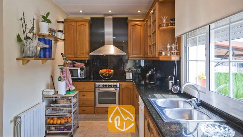 Ferienhäuser Costa Brava Spanien - Villa Mercedes Fenals - Küche mit Holzschränken, dunklen Granitarbeitsplatten, einem Edelstahlbackofen und einem Doppelwaschbecken unter einem Fenster.