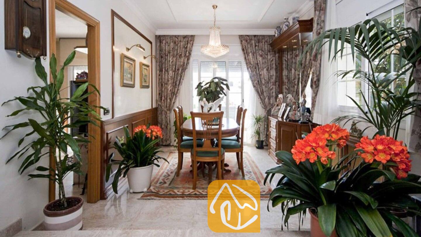 Vakantievilla's Costa Brava Spanje - Villa Mercedes Fenals - Interieur van de eetkamer met een houten tafel en stoelen, een kristallen kroonluchter, gordijnen met motief en potplanten die het uitzicht omlijsten bij Villa Mercedes Fenals.