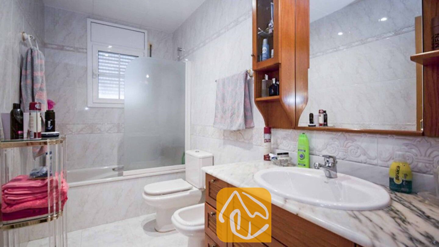Vakantievilla's Costa Brava Spanje - Villa Mercedes Fenals - Getegeld badkamer met een houten badkamermeubel, een wit marmeren wastafelblad, een ligbad met glazen wand en een hangende houten kast met spiegel.