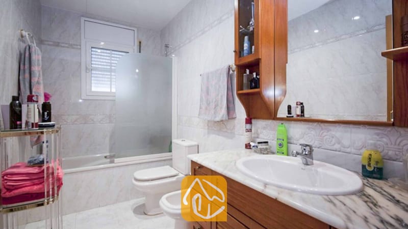Ferienhäuser Costa Brava Spanien - Villa Mercedes Fenals - Gefliestes Badezimmer mit Waschtisch aus Holz, weißer Marmorarbeitsplatte, Badewanne mit Glasabtrennung sowie einem wandmontierten Hängeschrank aus Holz mit Spiegel.