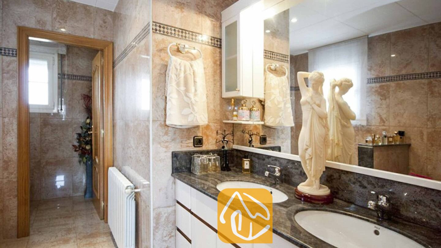 Vakantievilla's Costa Brava Spanje - Villa Mercedes Fenals - Badkamer voorzien van beige marmeren tegelwanden, twee witte wastafels op een donker granieten aanrechtblad, en een decoratief klassiek vrouwenstandbeeld.