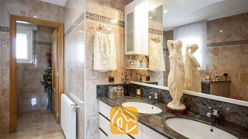 Ferienhäuser Costa Brava Spanien - Villa Mercedes Fenals - Badezimmer mit beige marmorierten Fliesenwänden, zwei weißen Waschbecken in einer dunklen Granit-Waschtischplatte und einer dekorativen klassischen Frauenstatue.