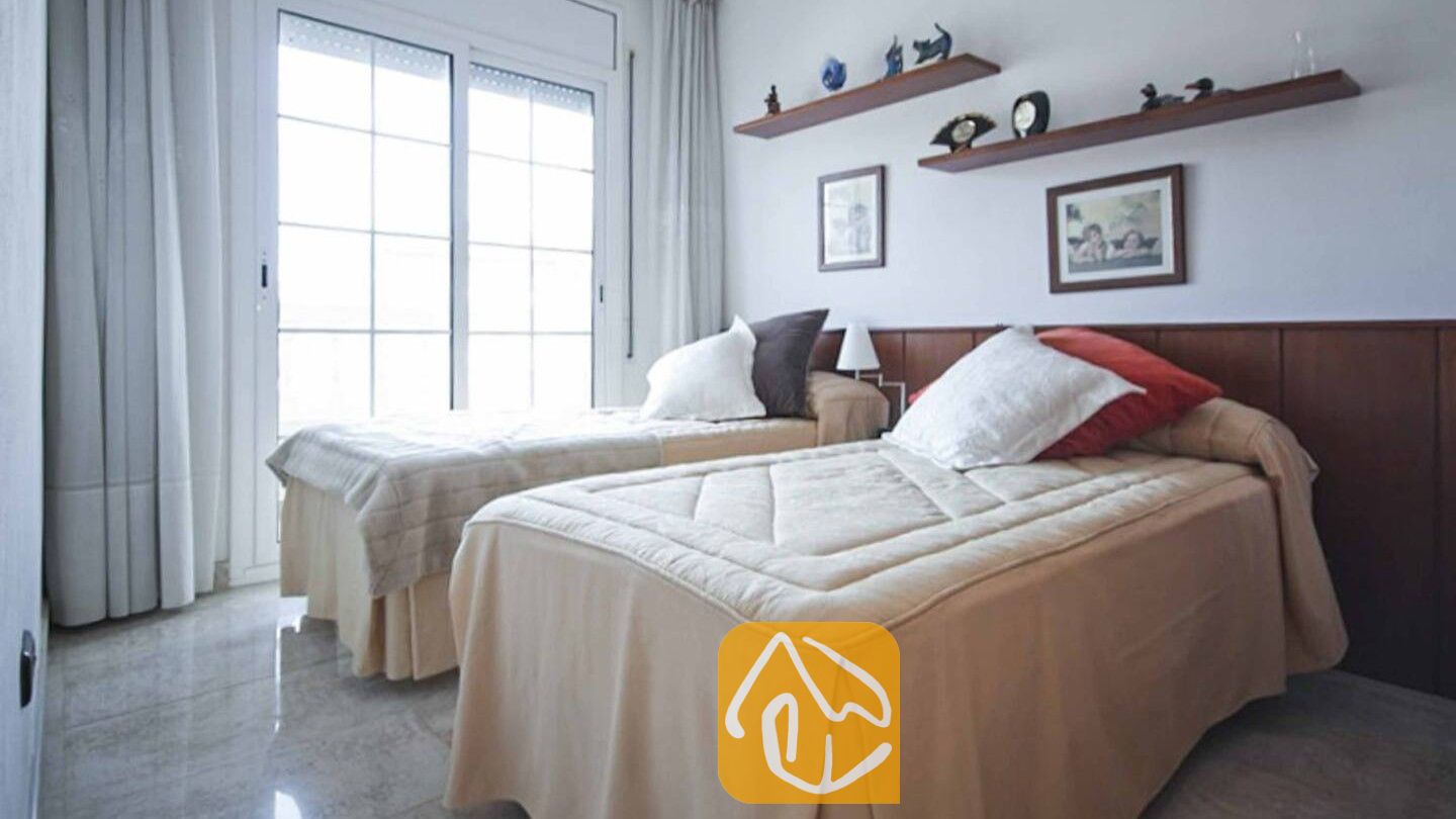 Vakantievilla's Costa Brava Spanje - Villa Mercedes Fenals - Slaapkamer met twee eenpersoonsbedden voorzien van beige spreien, witte en rode kussens, een houten hoofdbord en een grote schuifpui met witte gordijnen.