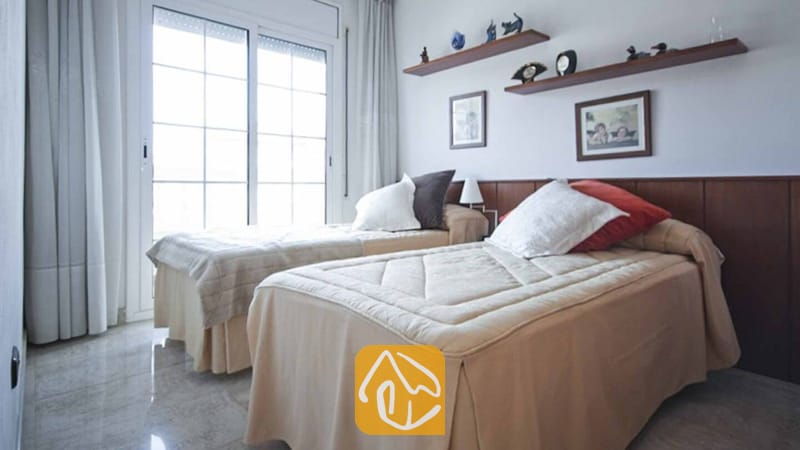 Ferienhäuser Costa Brava Spanien - Villa Mercedes Fenals - Schlafzimmer mit zwei Einzelbetten, bezogen mit beigen Tagesdecken, weißen und roten Kissen, einem Betthaupt aus Holz sowie einer großen Schiebetür aus Glas mit weißen Vorhängen.
